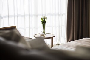 Ein Hotelzimmer mit einem Bett, einem Nachttisch mit Blumenvase und durchsichtigen Vorhängen.