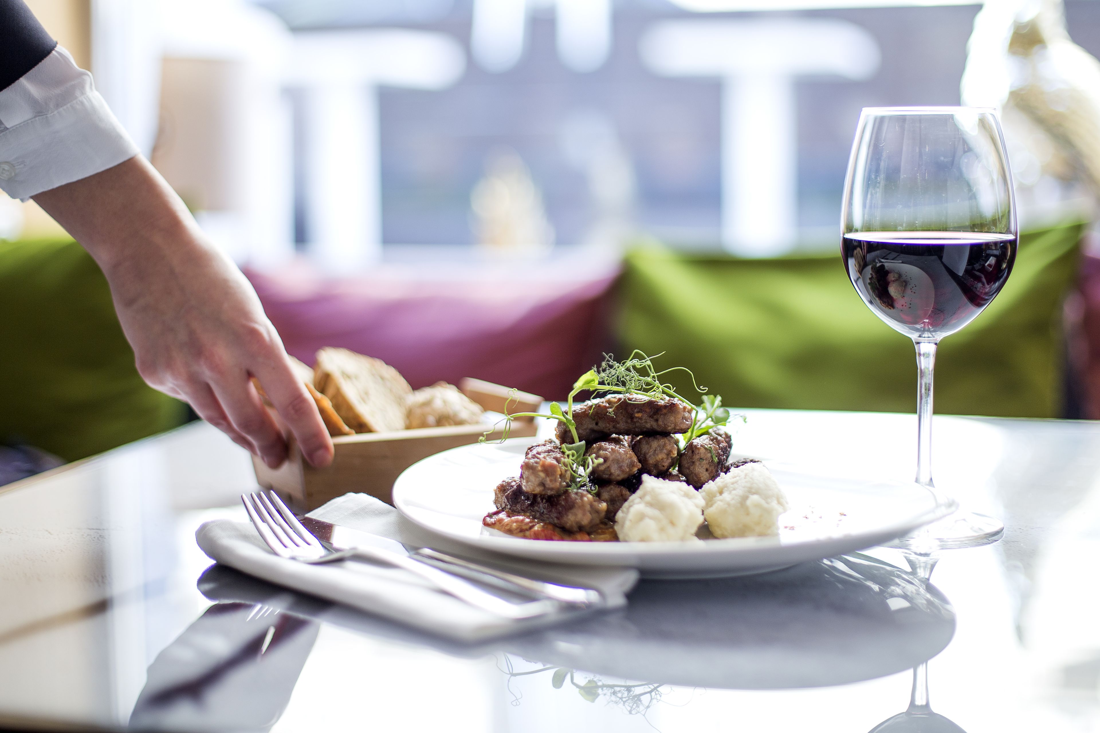 A dining table with a plate of food, a wine glass, and a hand reaching for a piece of bread.