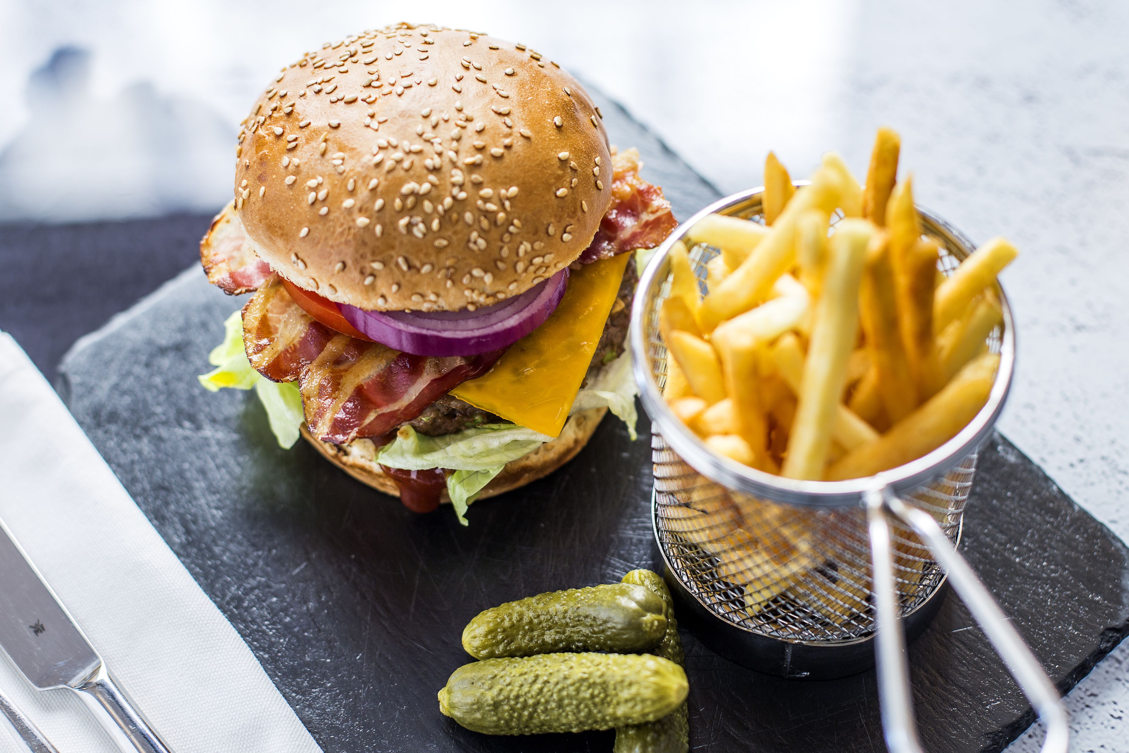 A burger with bacon, cheese, and lettuce on a sesame seed bun, accompanied by French fries and pickles on a slate serving board.