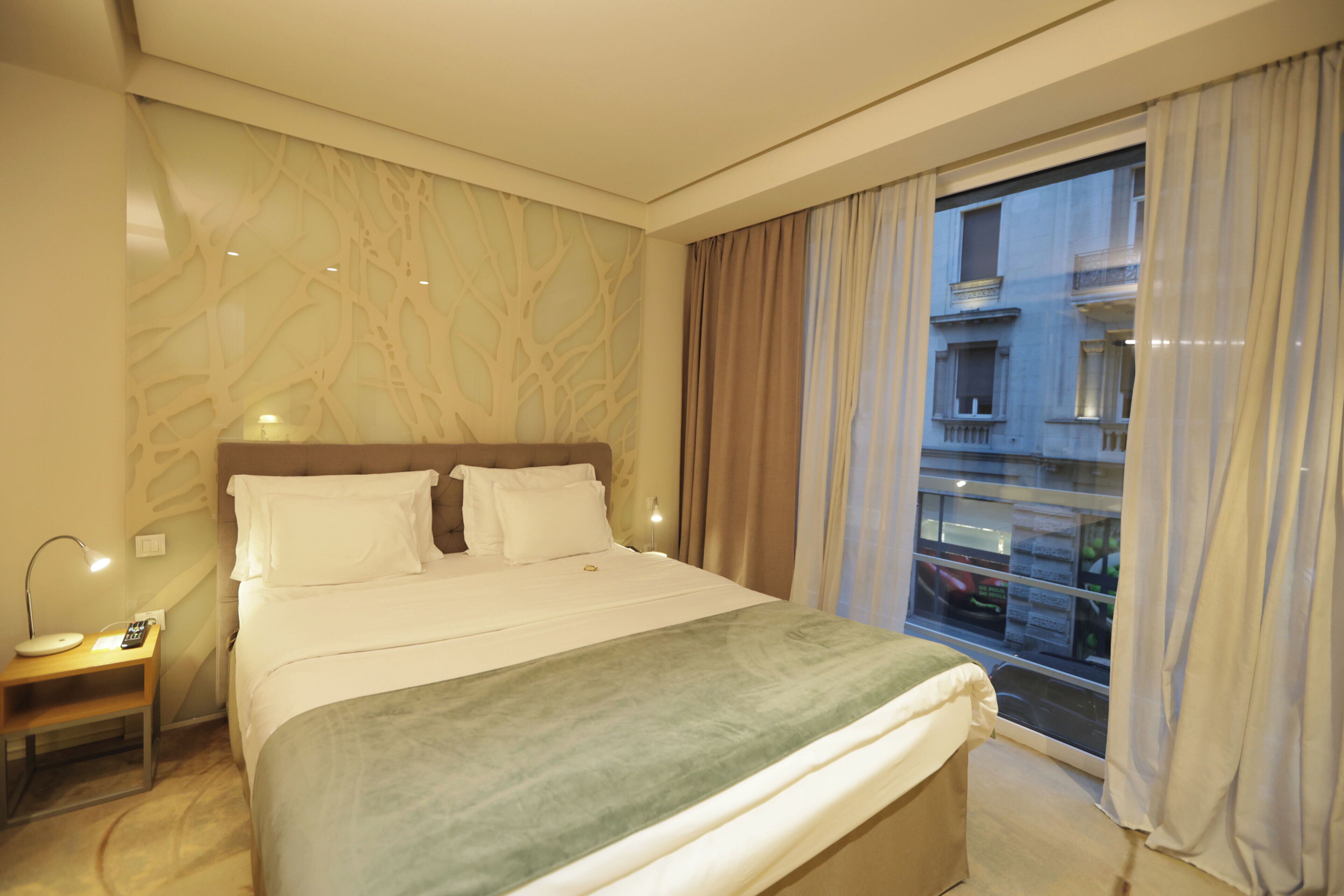 Habitación de hotel con una cama grande, una mesita de noche con lámpara y vistas a la ciudad a través de un ventanal amplio.