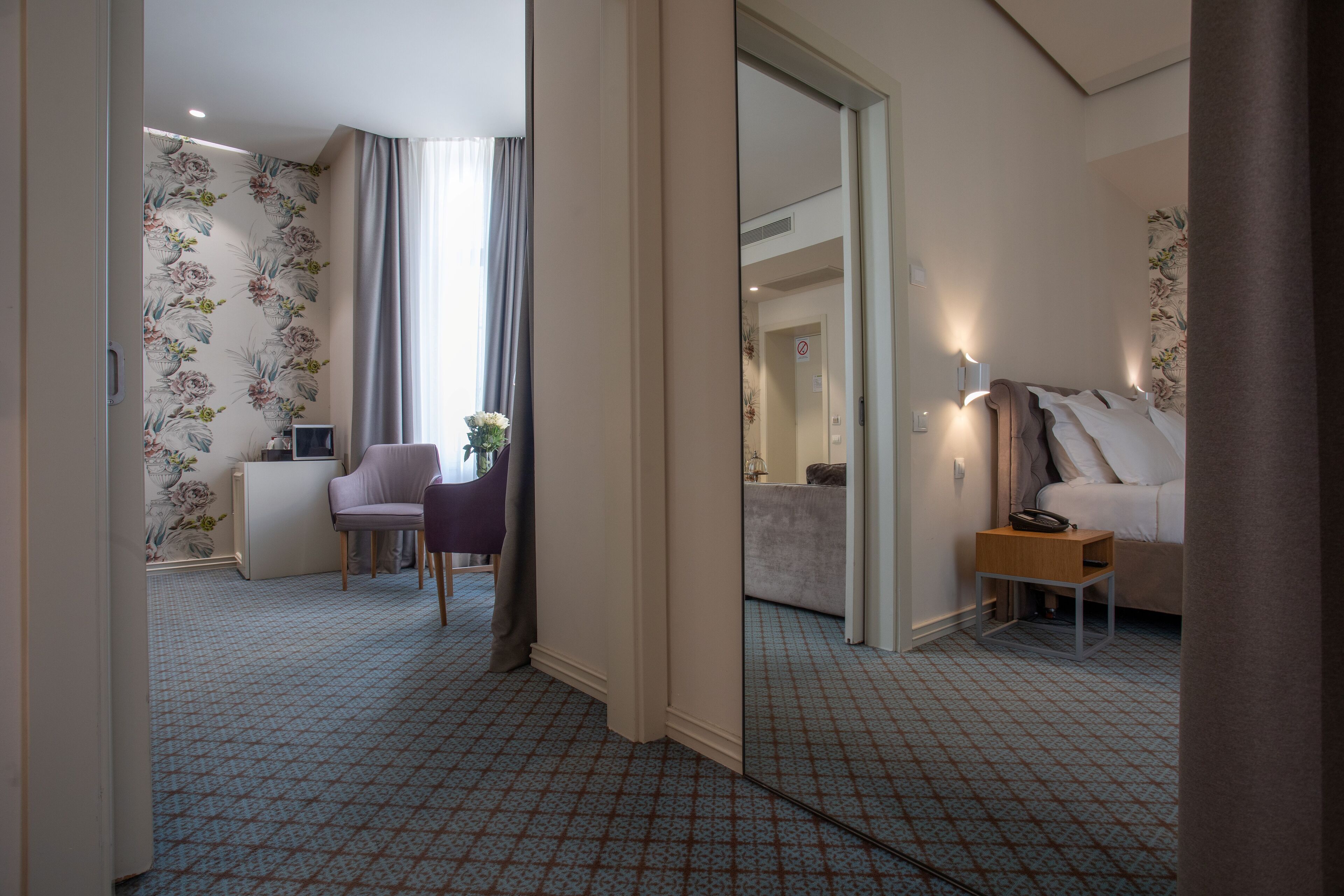 A hotel room with a patterned carpet, a bed, a bedside table with a lamp, a chair, and a desk with a computer.