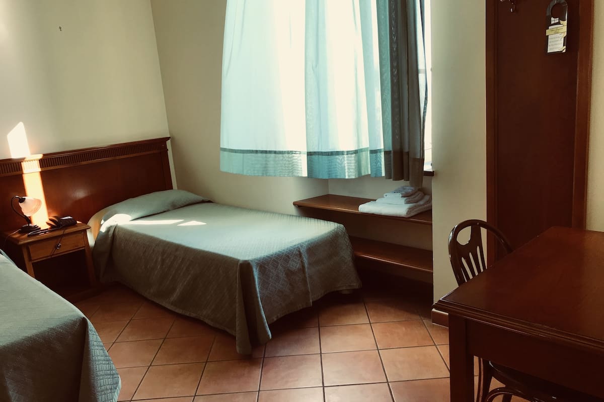 A small, clean room with two single beds, a wooden nightstand with a lamp, a small table, and a chair.