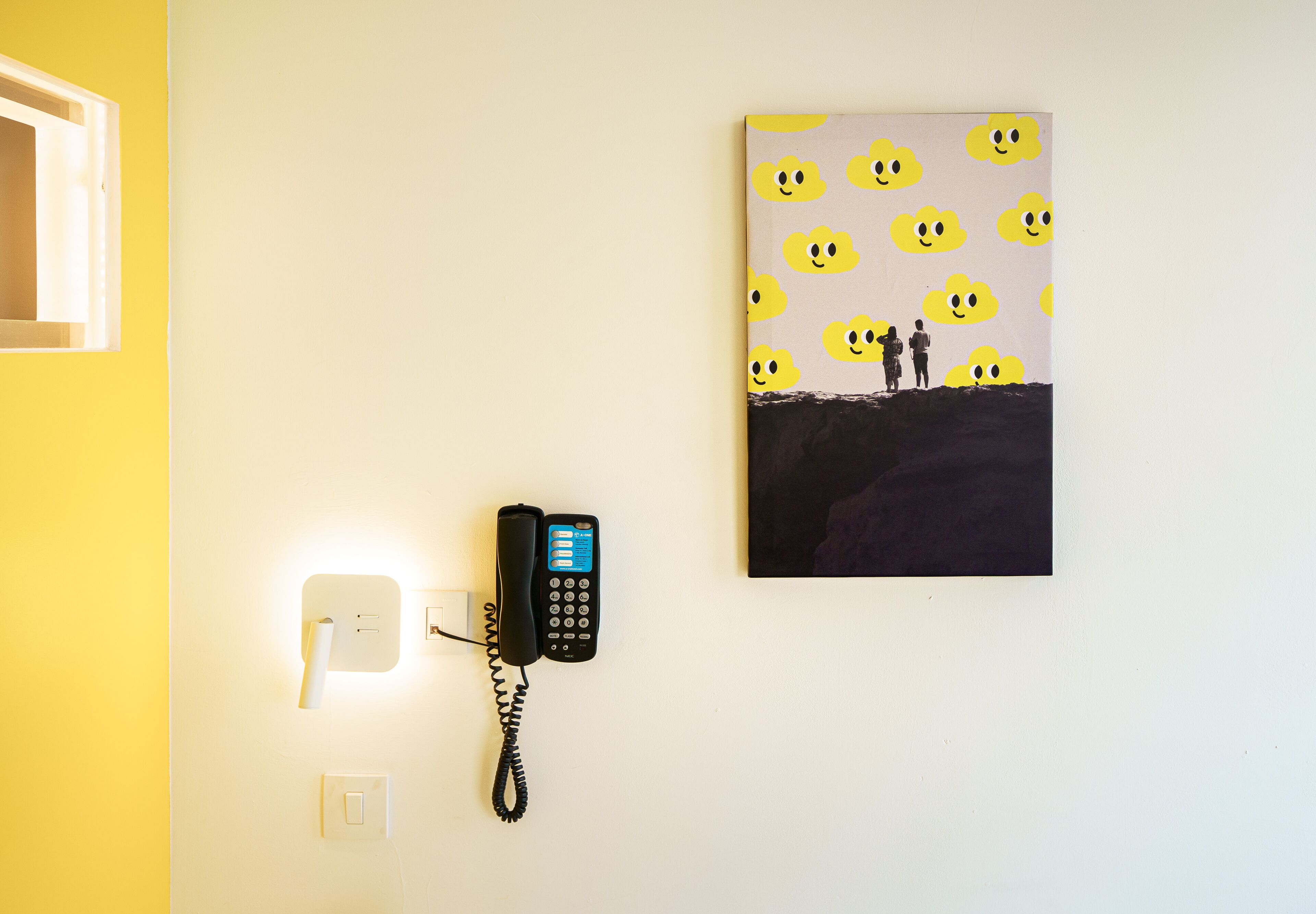 A wall-mounted telephone, a light switch, and a painting of two people standing on a cliff with a sky filled with smiley faces.