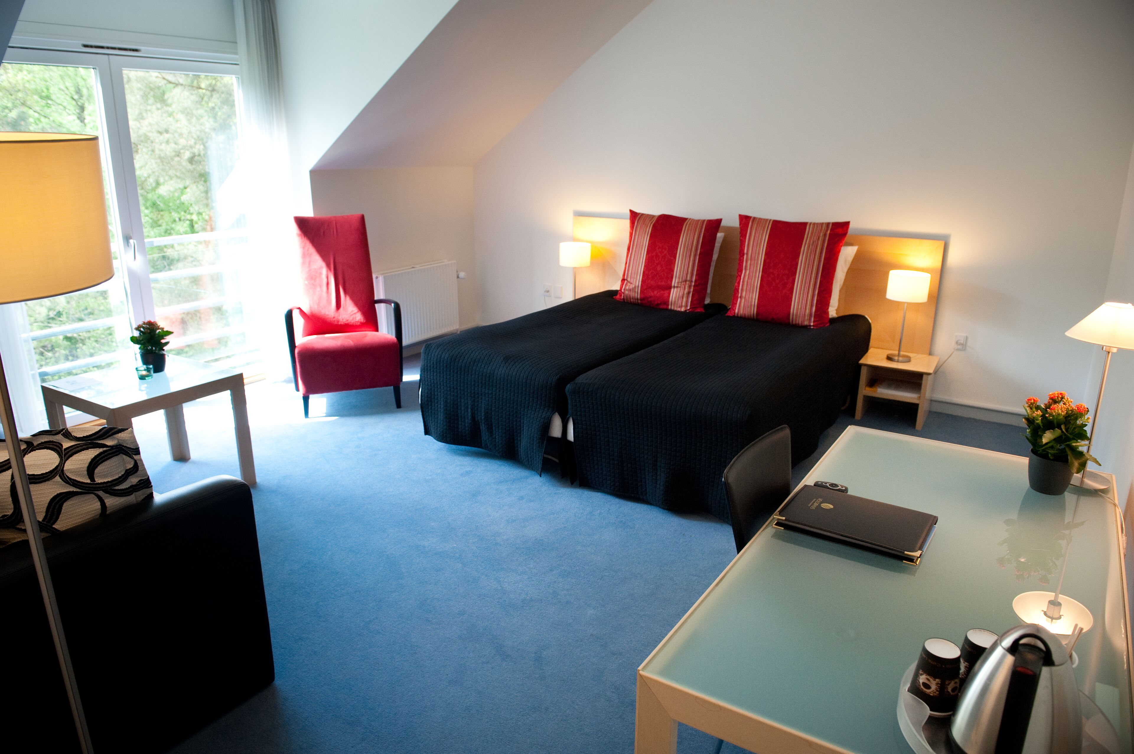 A hotel room with a bed, a desk, a chair, a lamp, and a view of greenery outside.