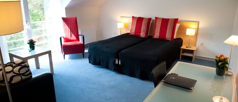 A hotel room with a bed, a desk, a chair, a lamp, and a view of greenery outside.