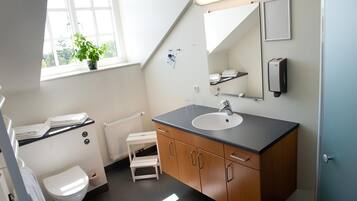 Un baño con inodoro blanco, lavamanos con espejo arriba, un taburete pequeño y una planta en maceta junto a la ventana.