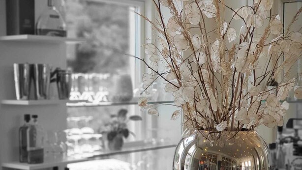 Une cuisine moderne avec une grande fenêtre, un vase contenant des branches séchées et un coin bar avec des bouteilles.