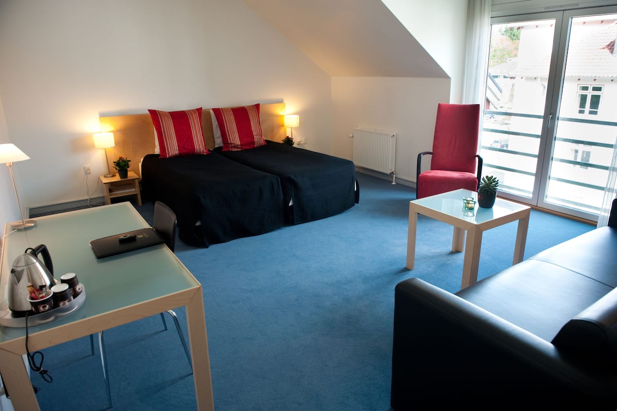 Une chambre d’hôtel avec un lit, une chaise rouge, une petite table avec une plante et un bureau avec une lampe et une bouilloire.