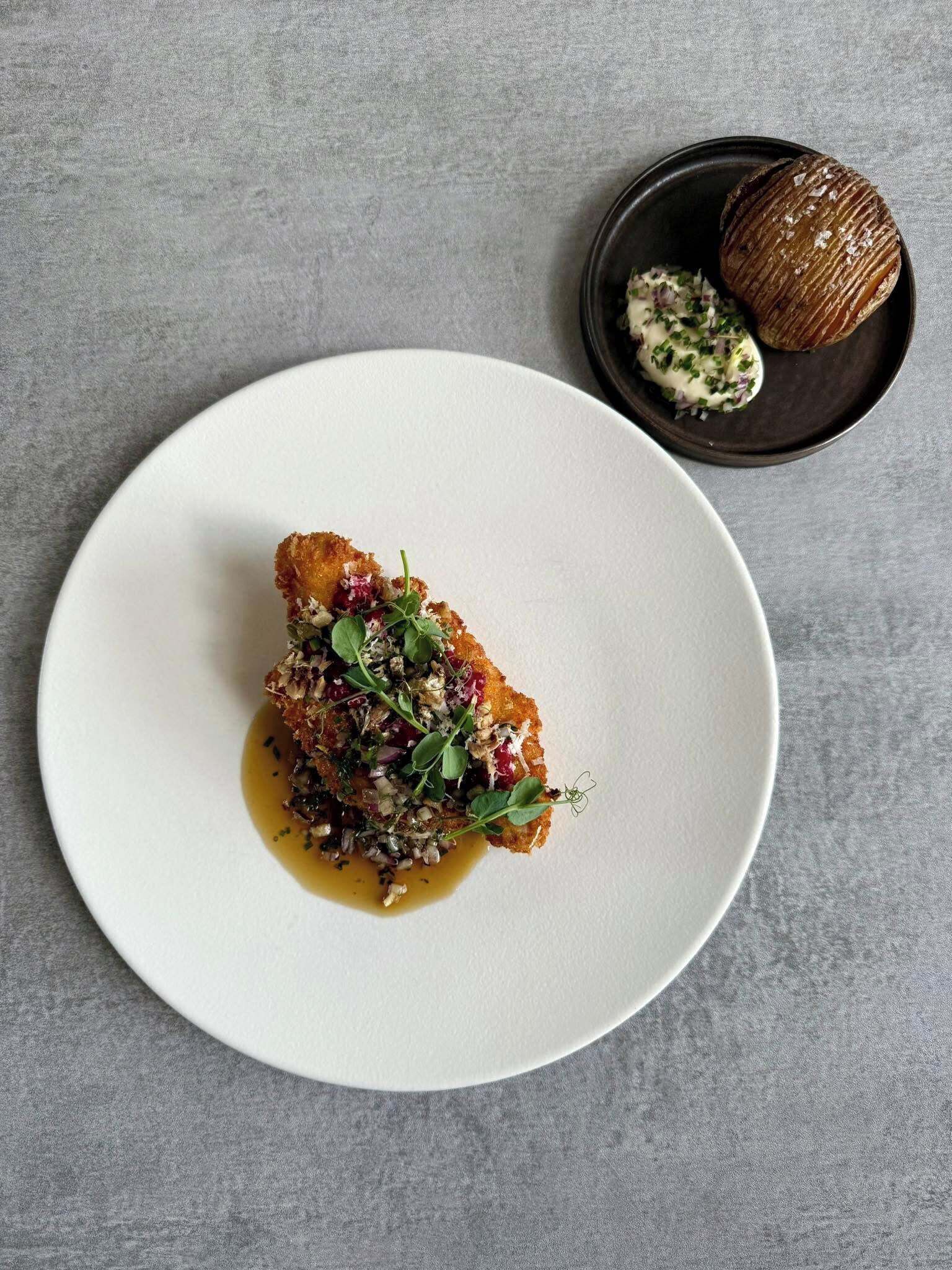 A white plate with a piece of food garnished with herbs and a sauce, and a small bowl with a dark-colored item and a creamy sauce.