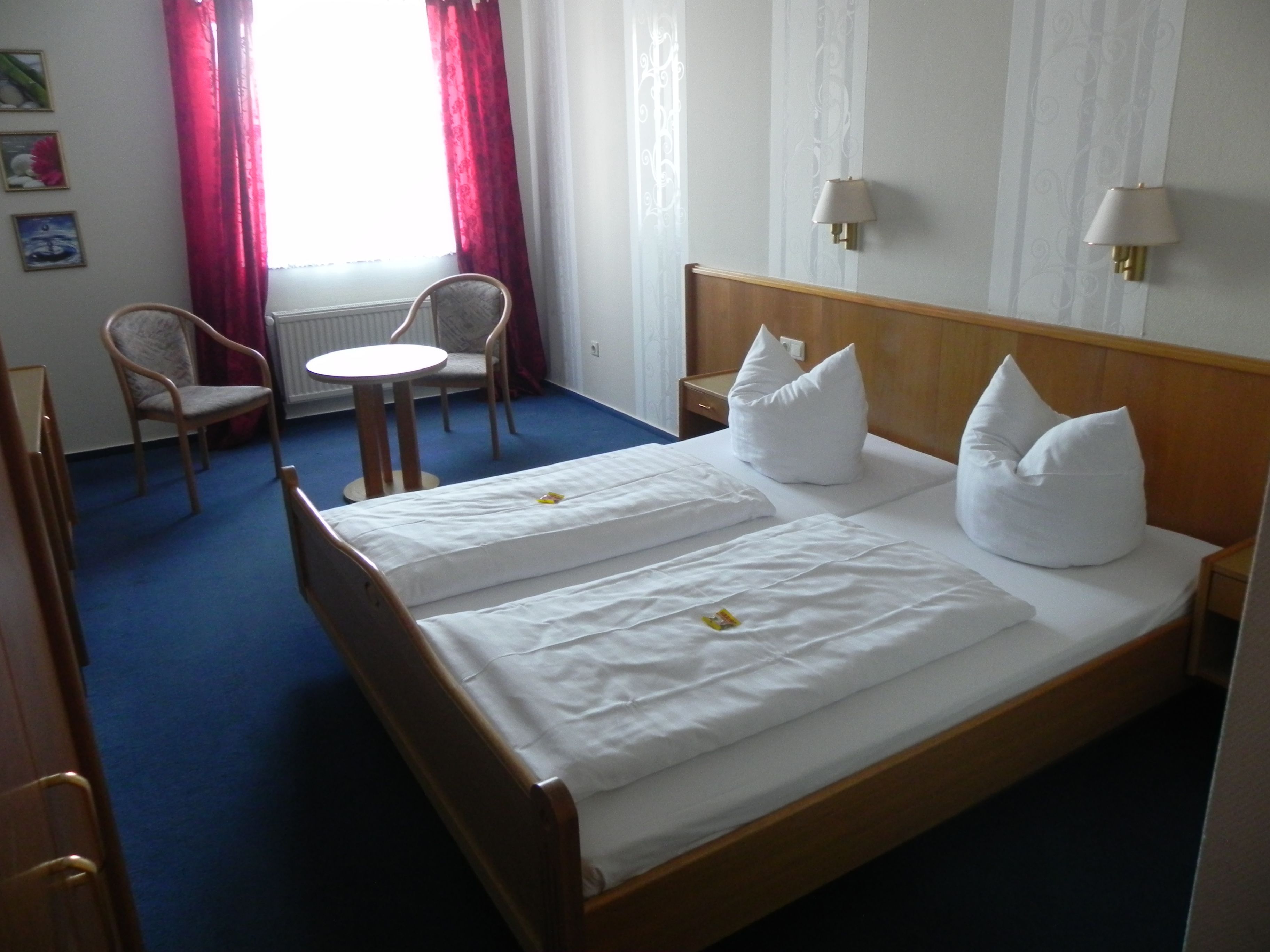 A hotel room with a double bed, two white pillows, a wooden headboard, a small round table with a chair, a desk, and a lamp.