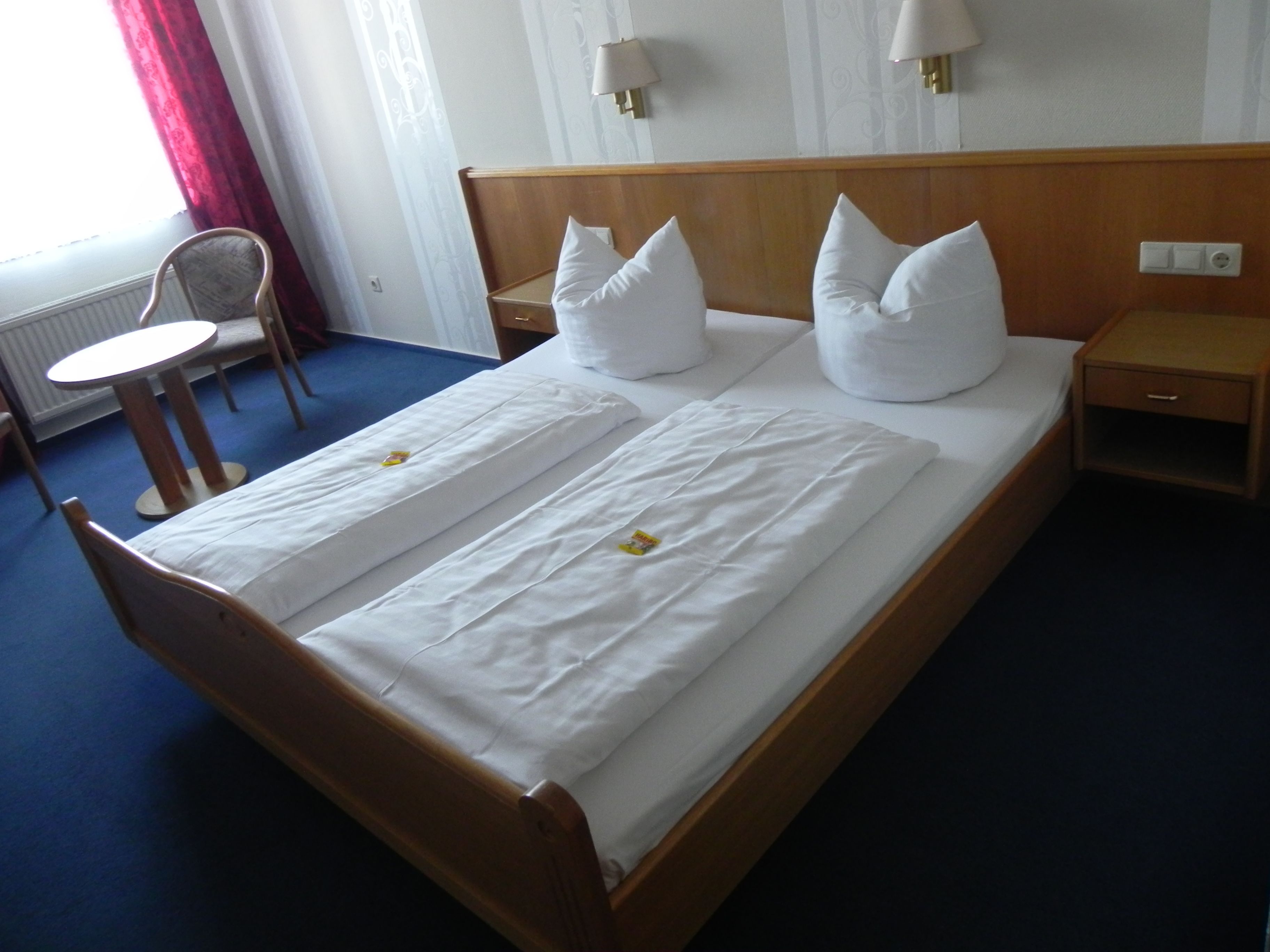 A neatly made bed with white linens and pillows, a wooden headboard, a bedside table, and a chair.