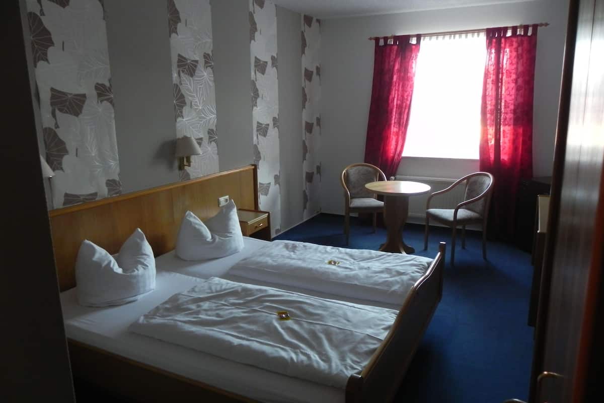 A hotel room with a bed, two pillows, a bedside table, a chair, a round table, and a window with red curtains.