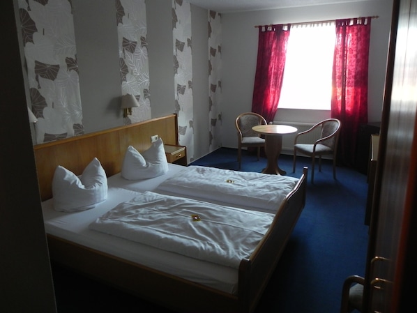A hotel room with a bed, two pillows, a bedside table, a chair, a round table, and a window with red curtains. - Hotel Zur Eldenburg (Luebz)