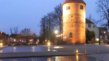A historic tower with a red roof, situated in a cobblestone square with a reflective water feature in the foreground.