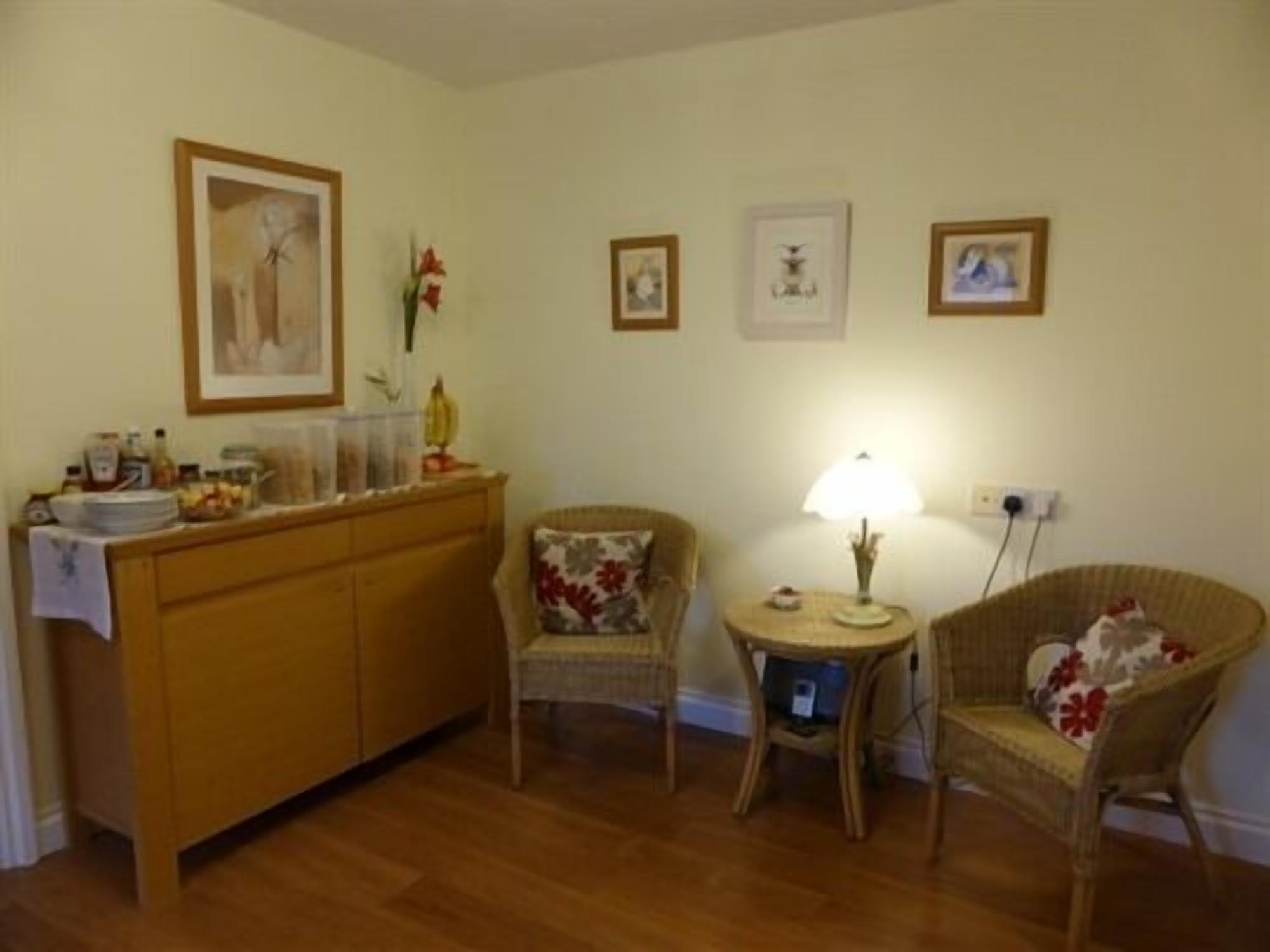 A cozy living room with a wooden cabinet, two wicker chairs, a small round table with a lamp, and framed pictures on the wall.