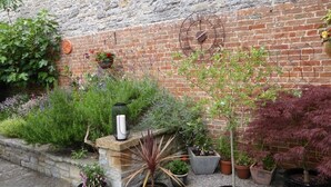 Un jardín con diversas plantas en macetas y un reloj de metal en una pared de ladrillo.