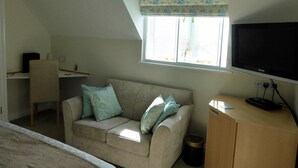 Una habitación con techo inclinado, un sofá color beige con cojines estampados, un televisor sobre un soporte de madera y una ventana con cortina floral.