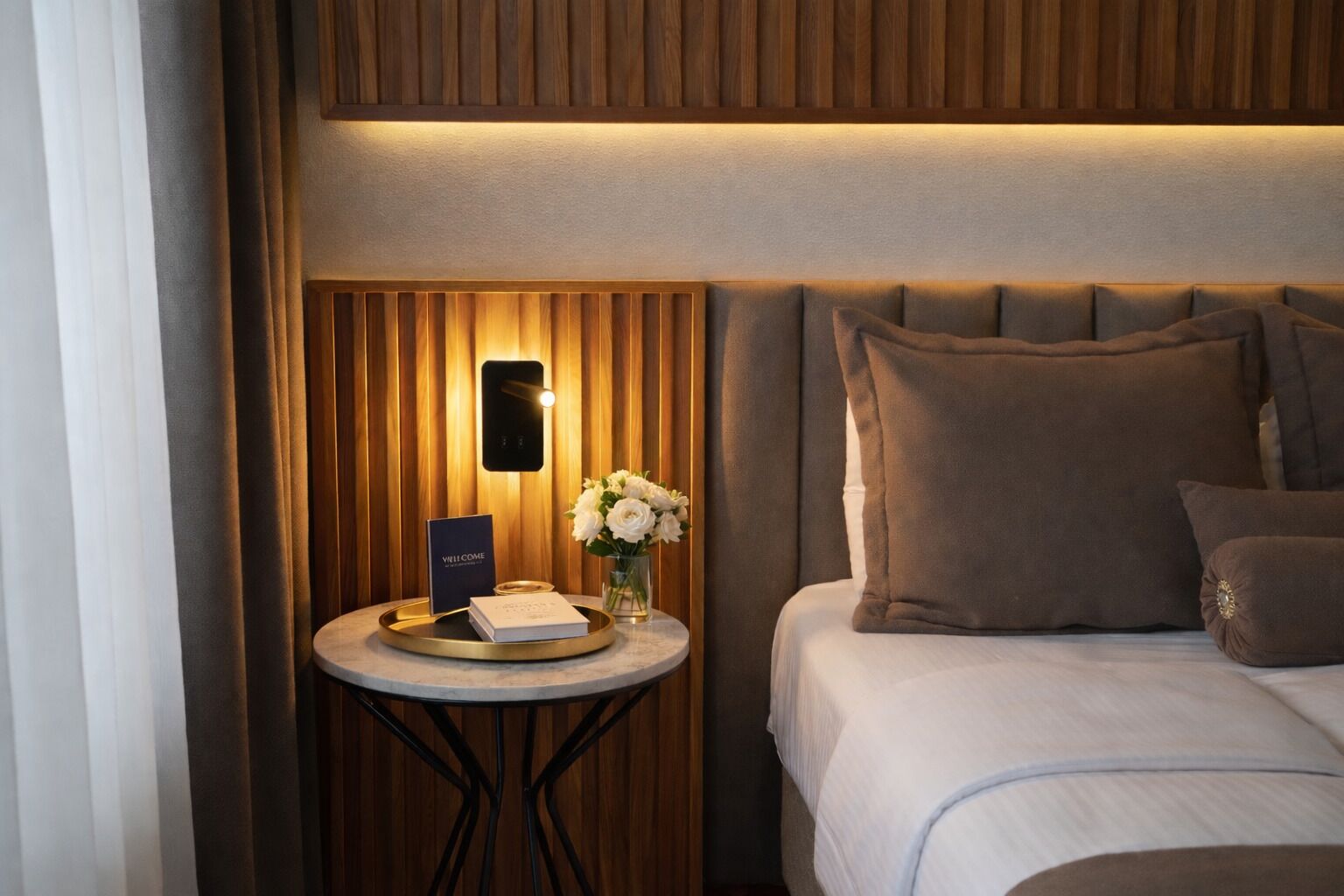 A hotel room with a bed, a bedside table with a lamp, a vase with flowers, and a sign that reads "WELCOME".