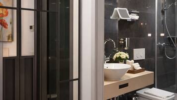 A modern bathroom with a glass door entrance, a large mirror, a sink with a vase of flowers, and a shower area.
