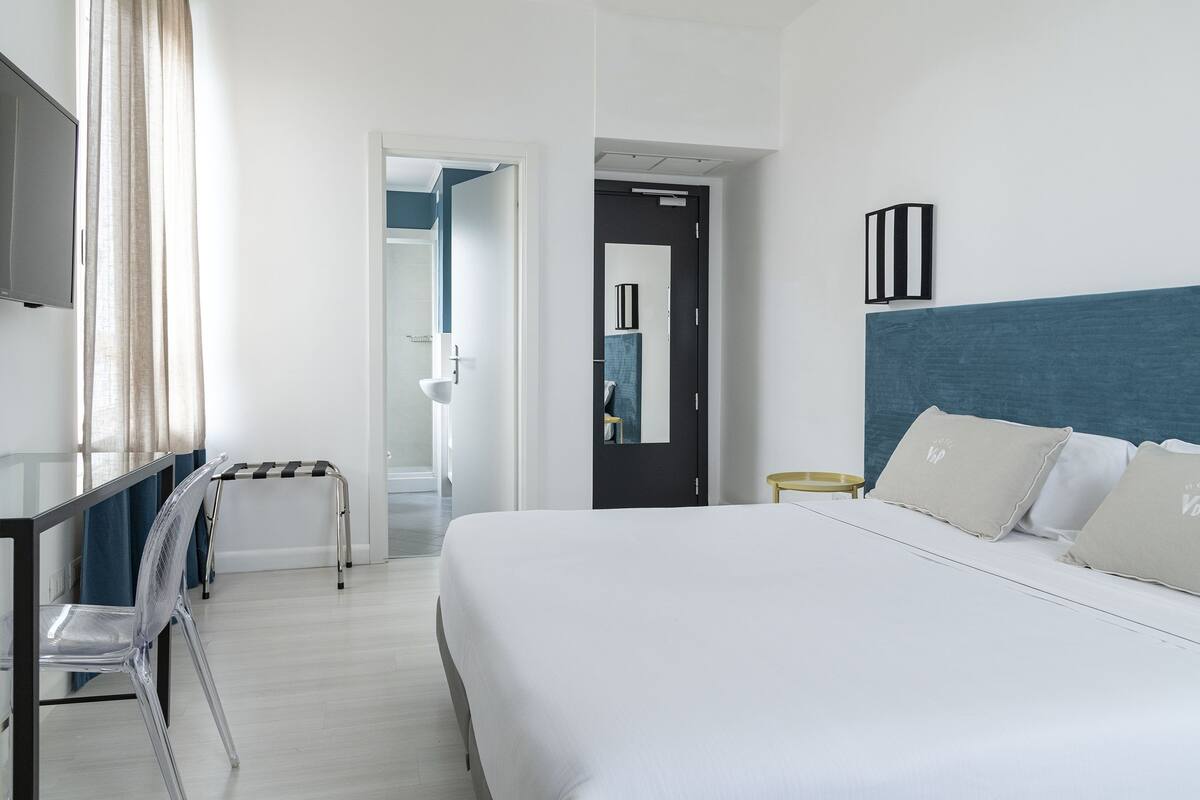 A hotel room with a large bed, a desk with a chair, a bedside table, and a TV mounted on the wall.