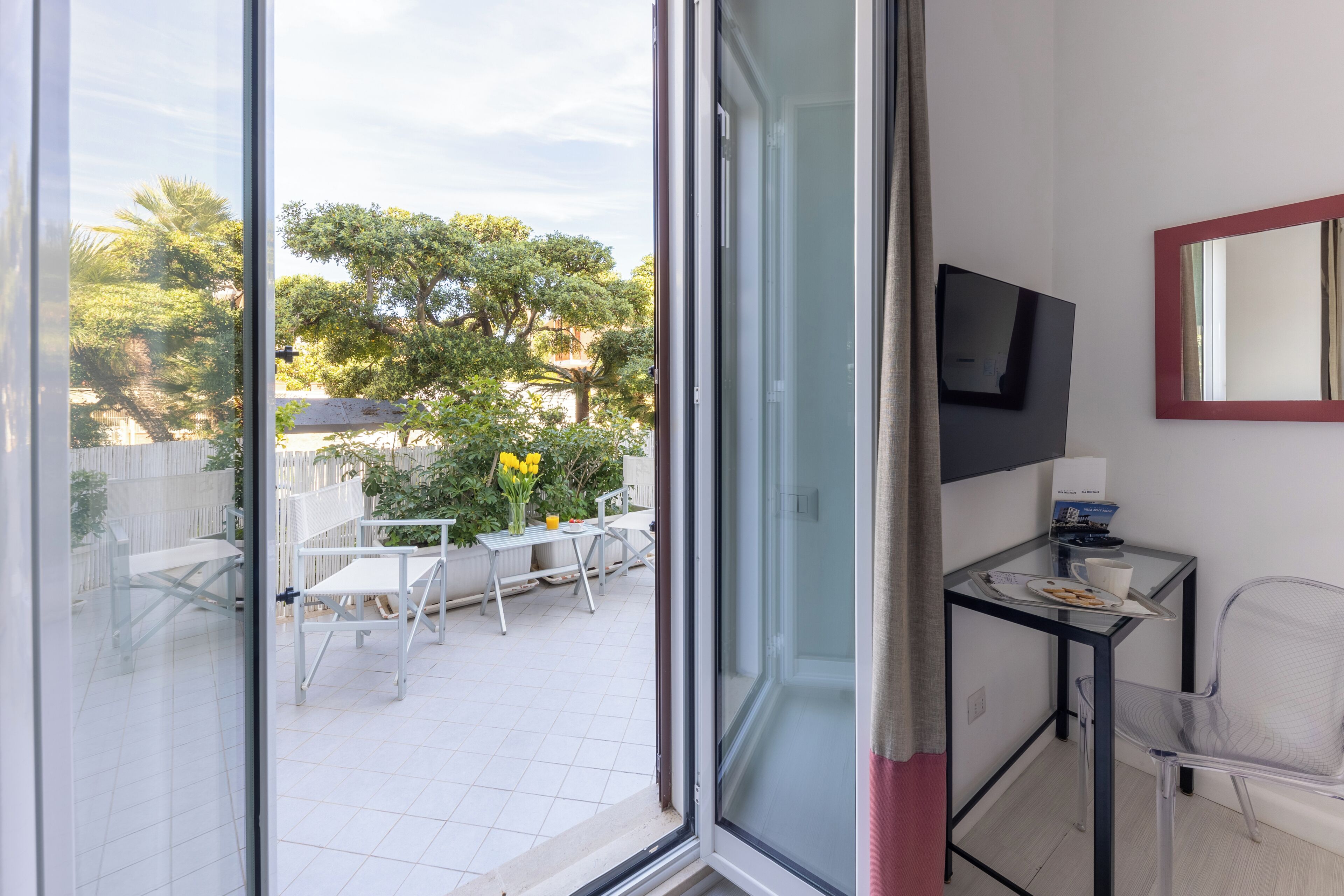 A room with a sliding glass door leading to an outdoor patio, a small table with a chair, a TV mounted on the wall, and a mirror.