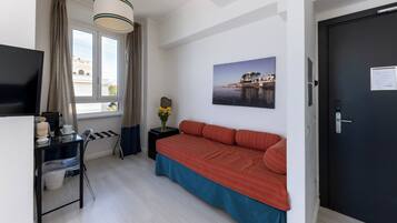 Ein Schlafzimmer mit einem Bett, einem Schreibtisch mit Stuhl, einem Fernseher, einem Fenster mit Vorhängen, einer Tür und einem Wandbild.