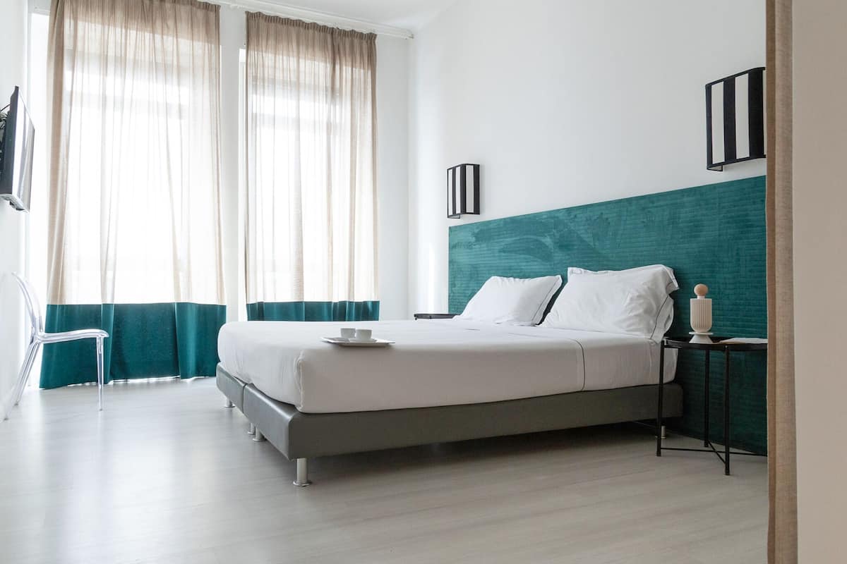 A modern hotel room with a bed, a side table, a chair, and a window with curtains.