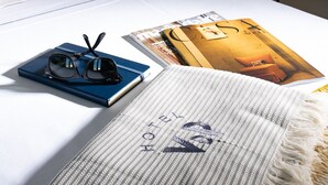Un lit bien fait, avec un oreiller portant l’inscription « HÔTEL VDP », une paire de lunettes, un magazine et un plaid rayé.