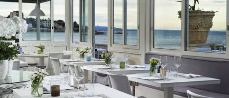 A restaurant with tables set for dining, large windows with a view of the sea, and a potted plant on the windowsill.
