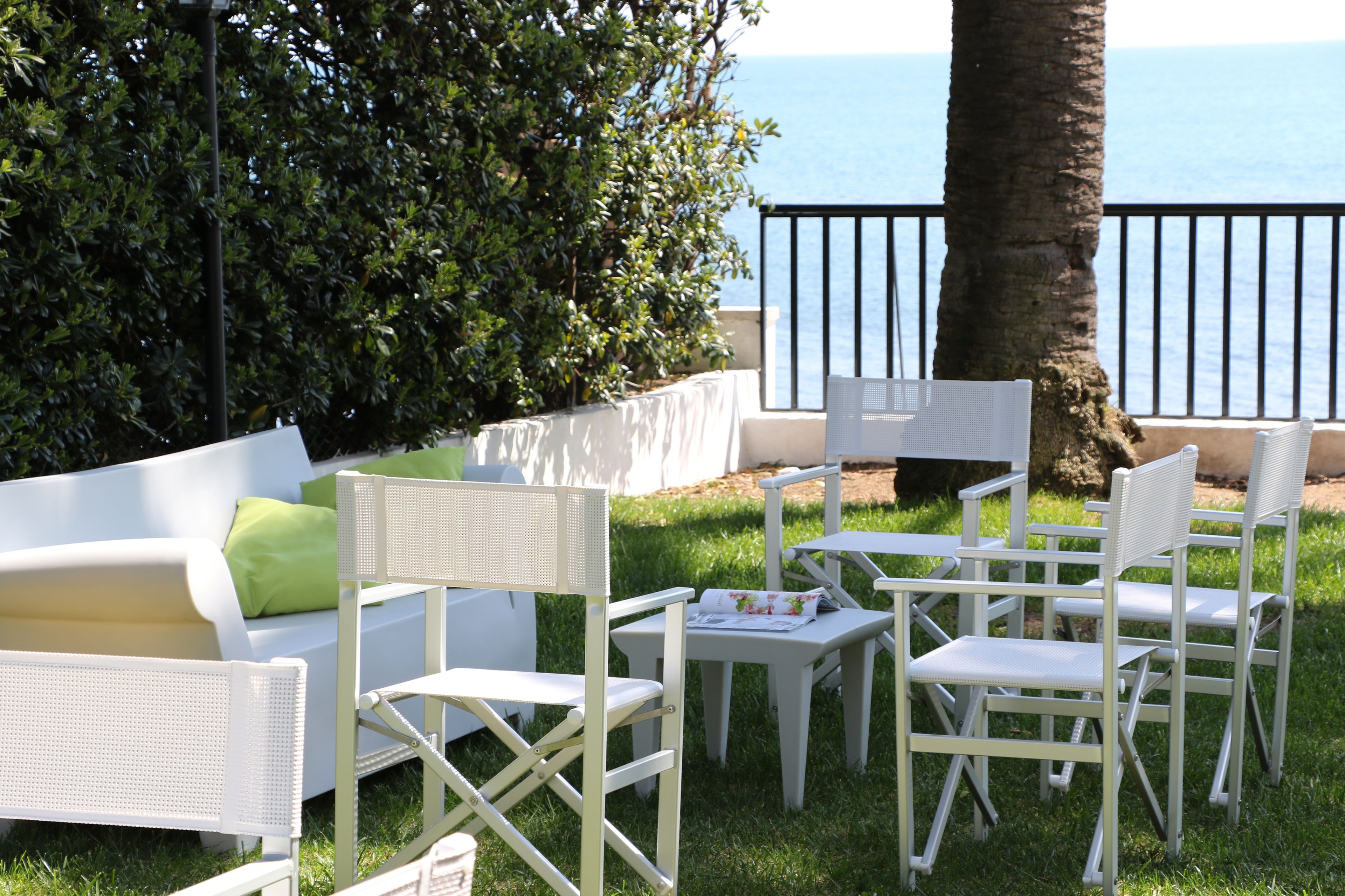A terrace with white furniture, including a sofa and chairs, set against a backdrop of a palm tree and a view of the sea.