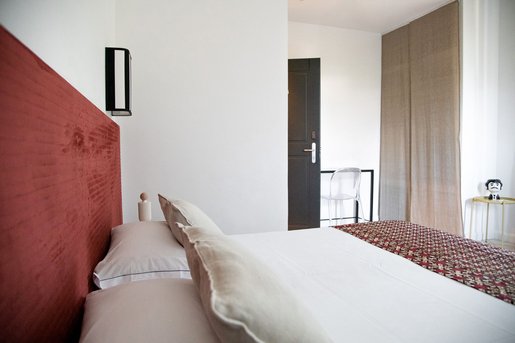 Habitación de hotel con una cama grande, una pared decorada en rojo, una mesita con una silla y una puerta que da a otra habitación.