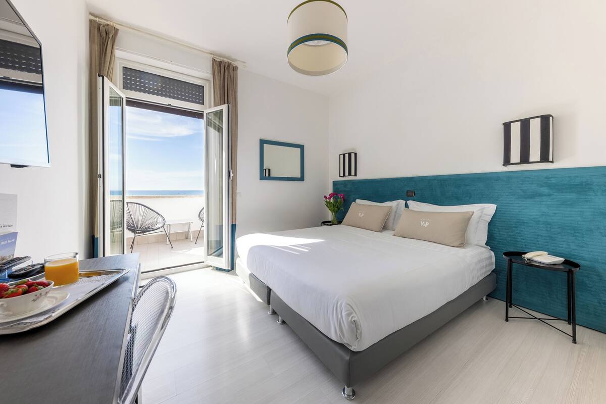 A hotel room with a bed, a desk with breakfast, a phone, and a view of the sea.