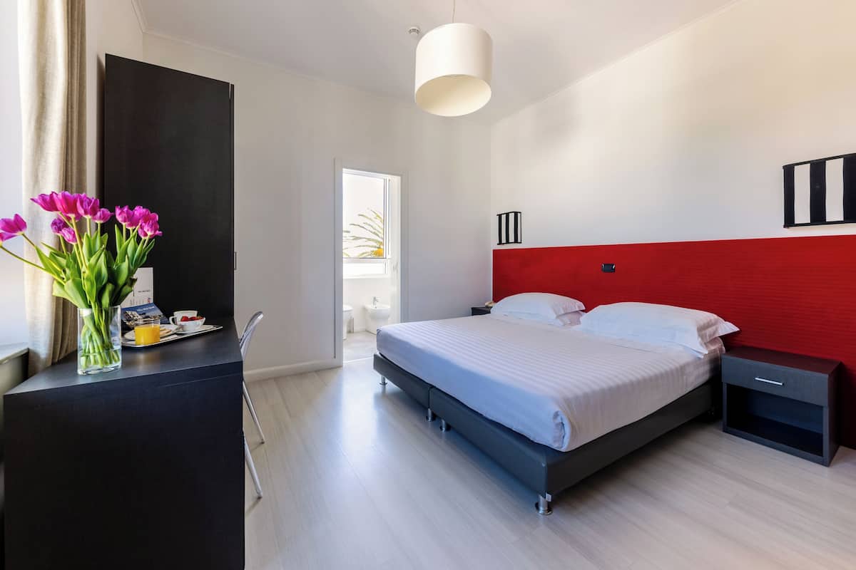 A modern bedroom with a bed, bedside tables, a desk with a vase of flowers, and a red accent wall.