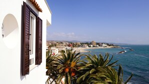 Un bâtiment situé sur la côte, doté d’un balcon donnant sur la mer, avec des palmiers au premier plan et une vue sur la plage et le paysage urbain en arrière-plan.
