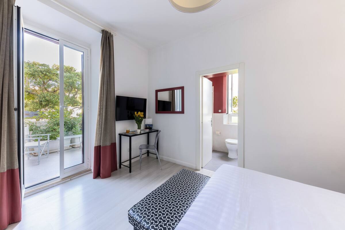 A hotel room with a bed, a desk with a chair, a TV, a mirror, and a bathroom door.