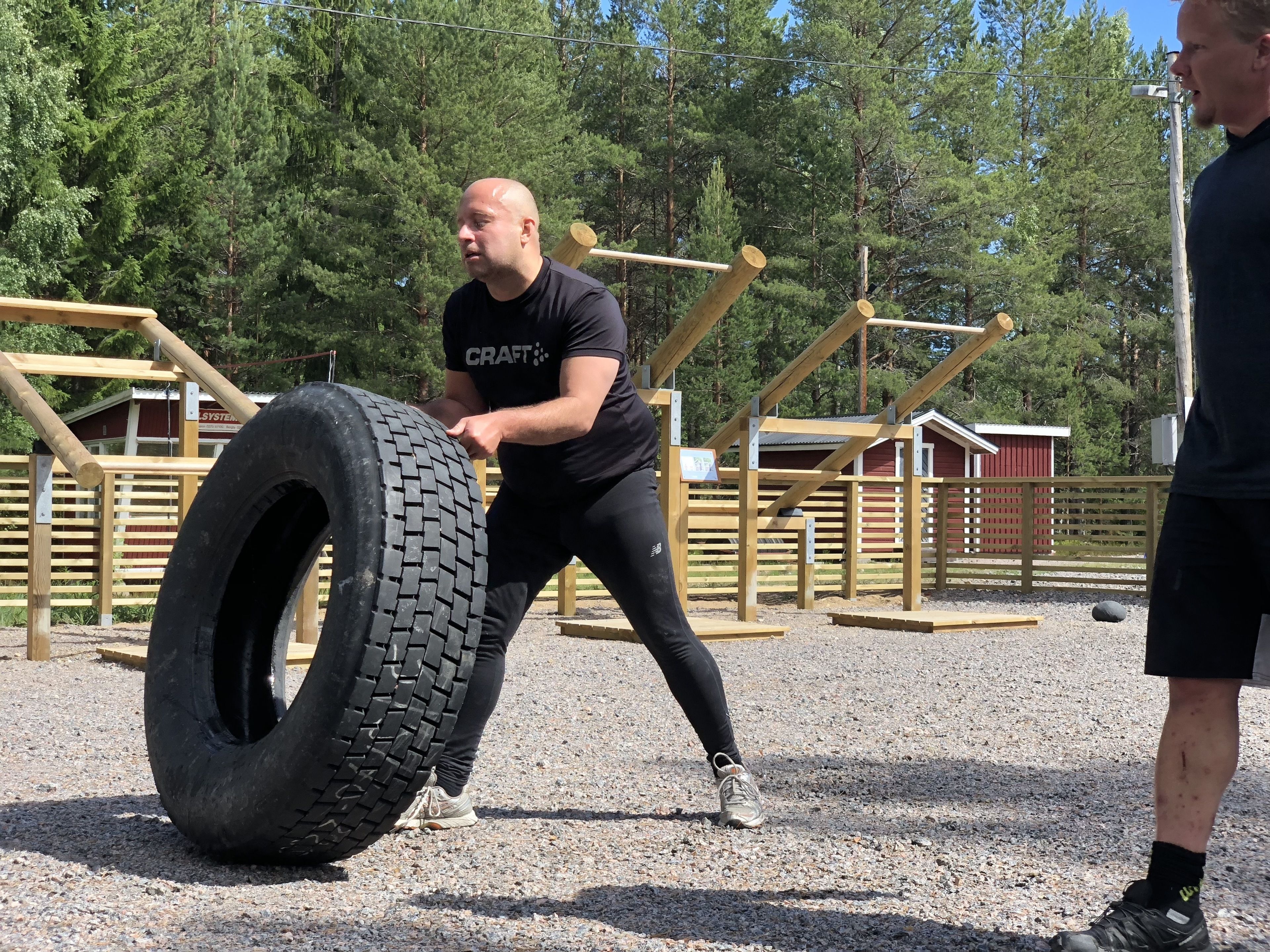 A person performing a tire flip exercise in an outdoor setting with wooden structures and trees in the background.