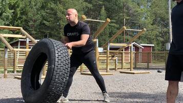 A person performing a tire flip exercise in an outdoor setting with wooden structures and trees in the background.