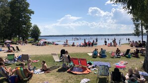 Escena de playa con gente relajándose en sillones de jardín y mantas, cielo despejado y un lago tranquilo al fondo.