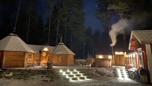 Una cabaña de madera con techo nevado, un pequeño porche con sillas y una chimenea visible que emite humo.