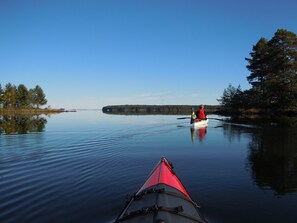 A calm lake with a red kayak in the foreground and a person in a red life jacket paddling a small boat in the background. - Hedenstugan (Bergby)