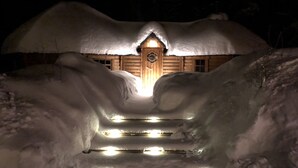 Una cabaña de madera cubierta de nieve con una puerta central y escalones que llevan a ella, iluminada por luces.
