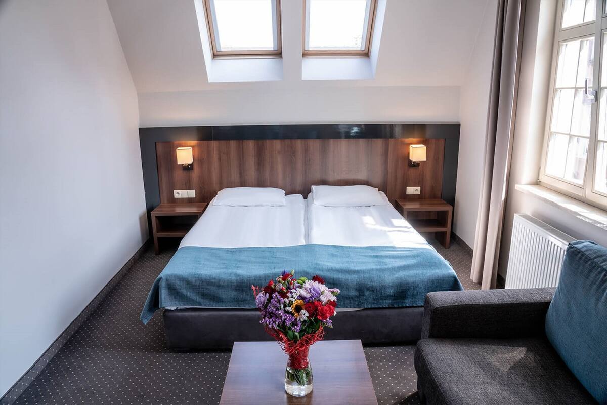 Une chambre d’hôtel comprenant un lit, deux tables de chevet, une commode, une fenêtre avec des rideaux, une petite table ornée d’un vase de fleurs et un canapé.