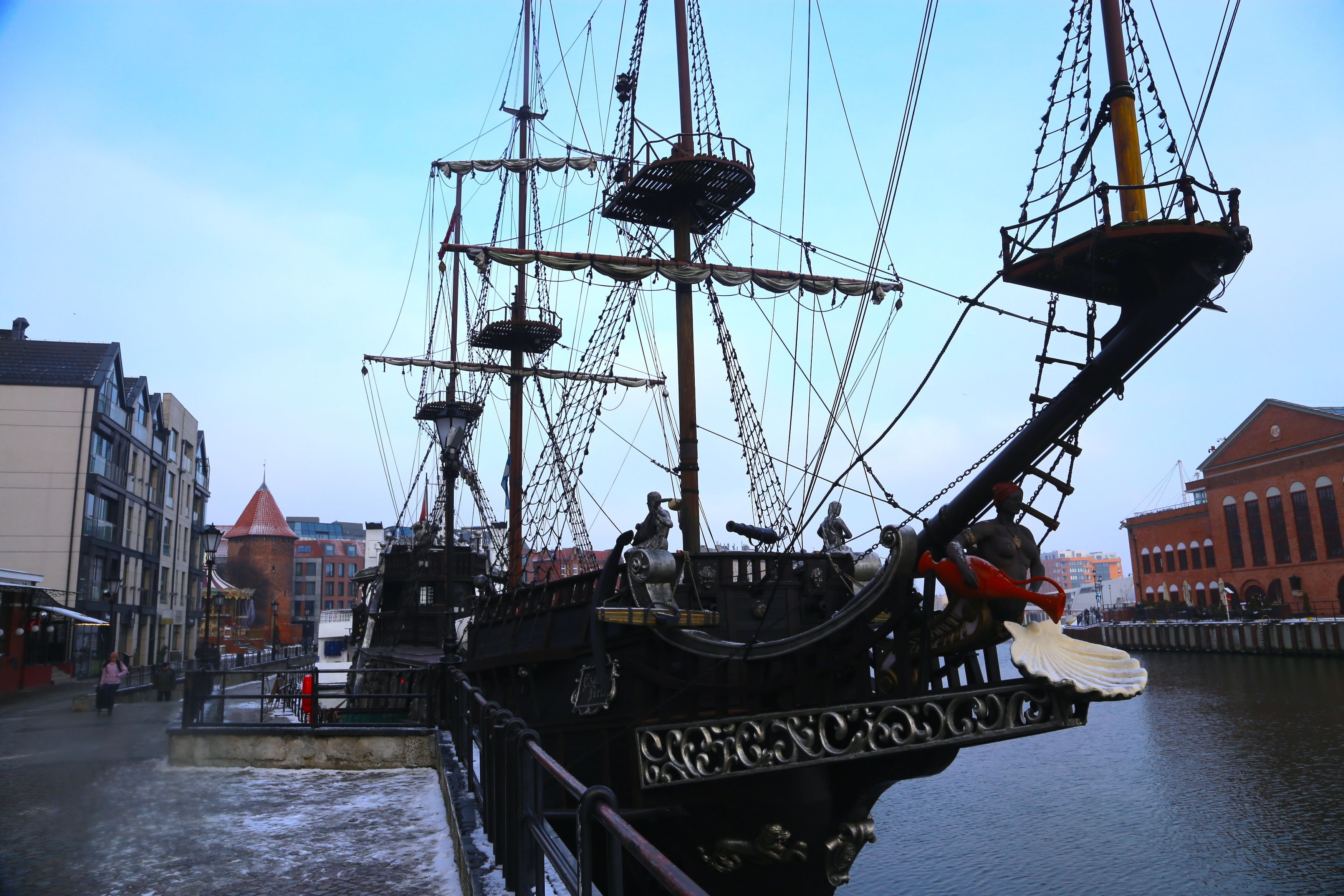 A tall ship docked at a pier with a cityscape in the background.