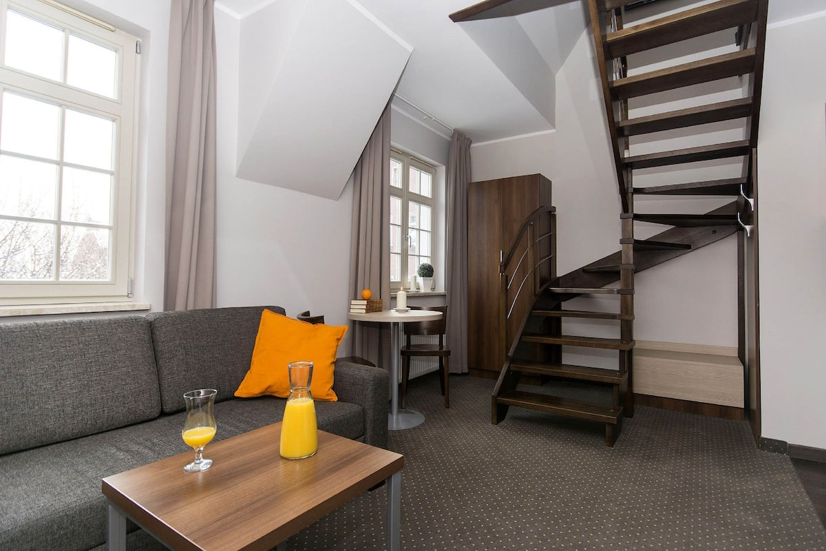 Un salon moderne avec un canapé gris, une table basse en bois sur laquelle se trouve un verre de jus d’orange, une petite table ronde avec une bougie blanche et un escalier en bois.