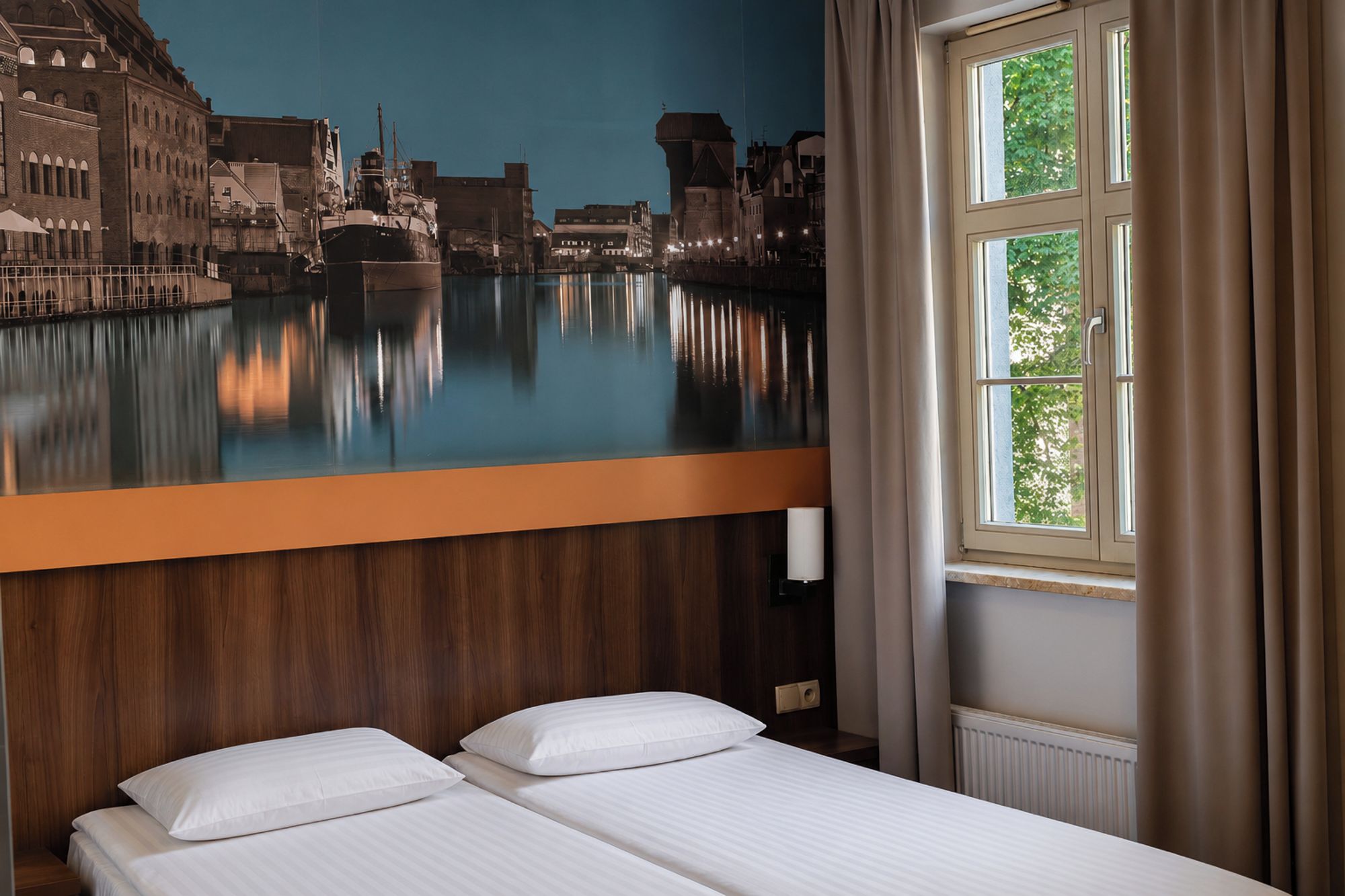 A hotel room with a bed, a window with curtains, and a mural of a cityscape on the wall.