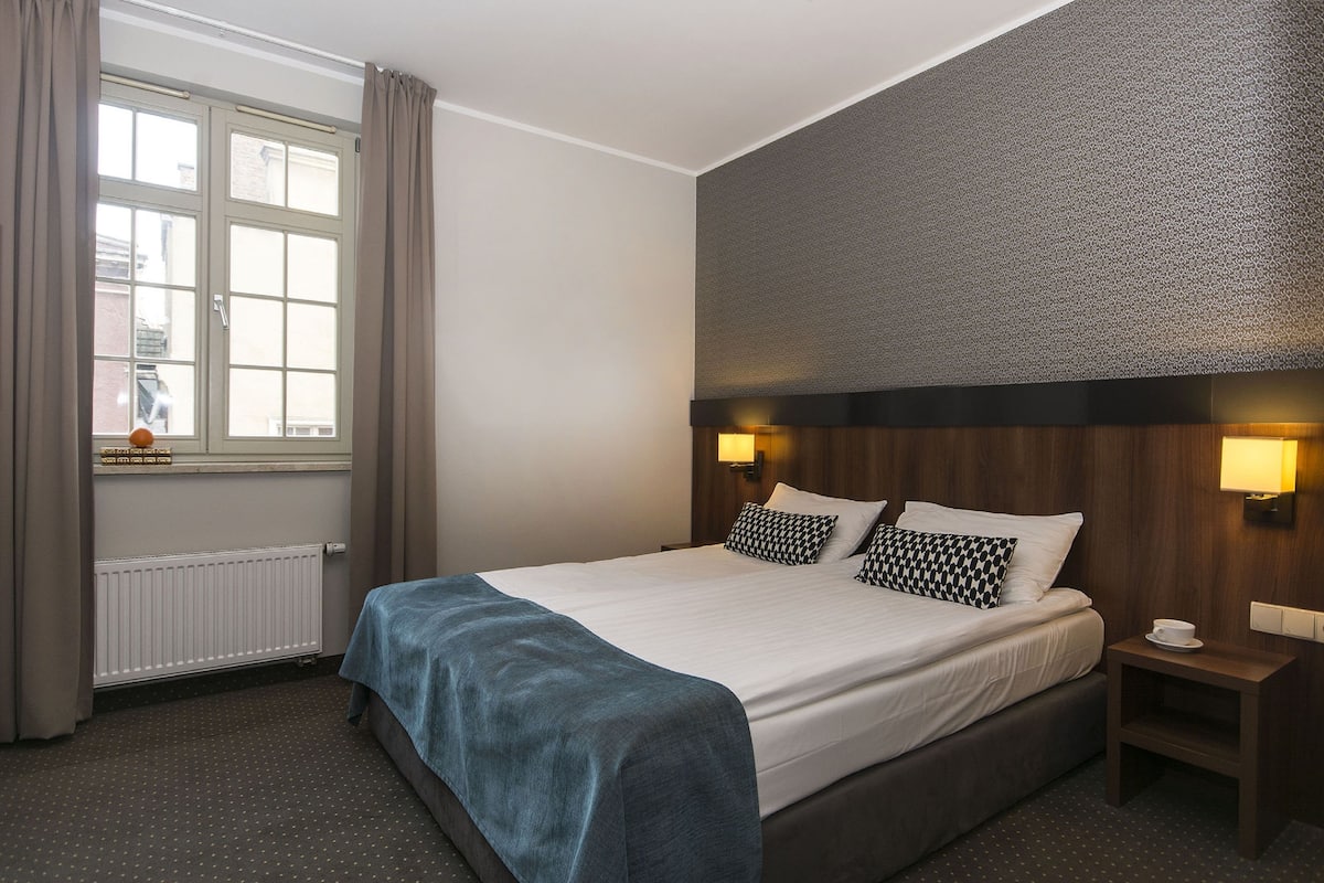 Une chambre d’hôtel avec un grand lit, une fenêtre avec des rideaux, une table de chevet avec une tasse et une lampe fixée au mur.