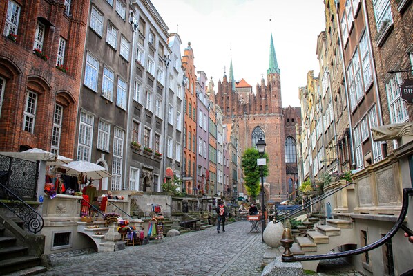 Una calle empedrada flanqueada por edificios históricos, la aguja de una iglesia y puestos de mercado al aire libre.