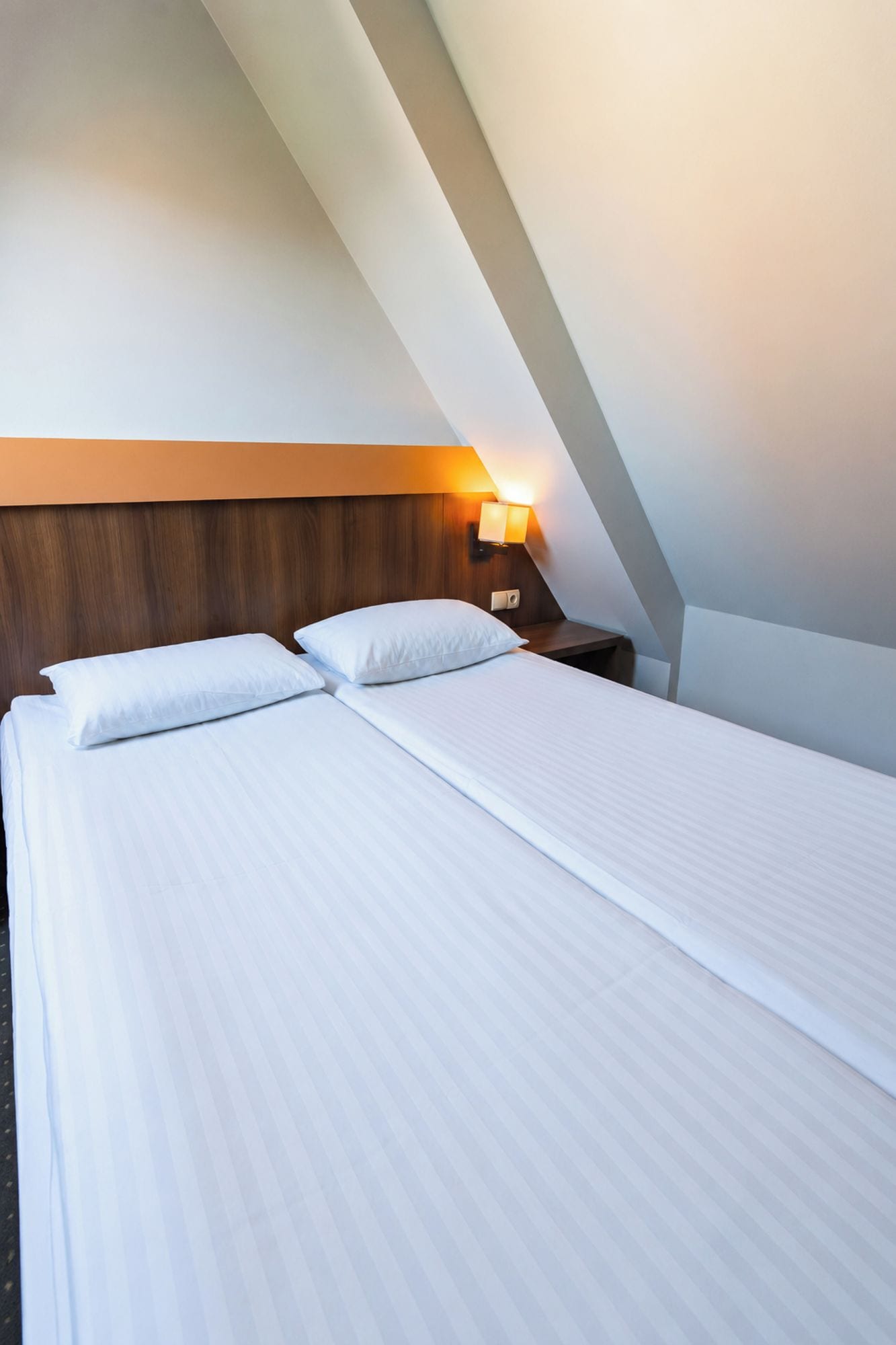 A neatly made bed with white linens in a room with a sloped ceiling and a wall-mounted nightstand with a lamp.