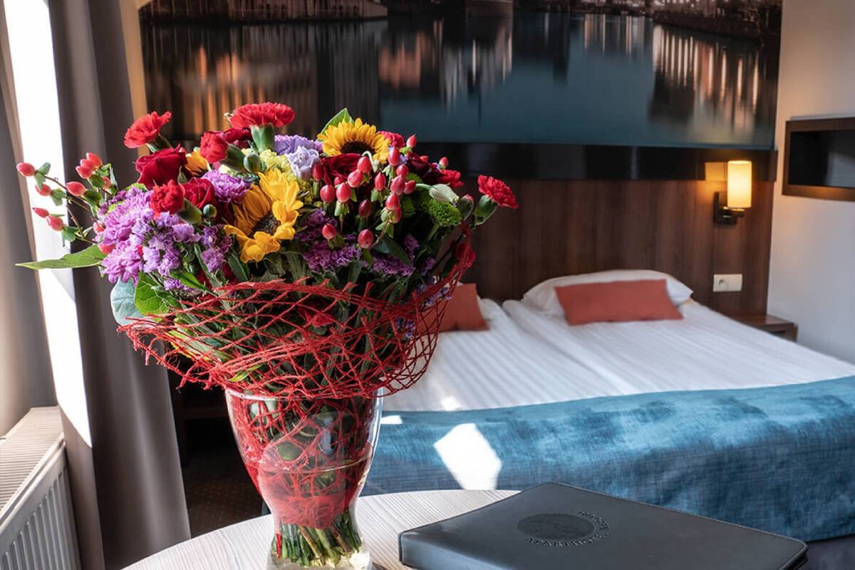 Une chambre d’hôtel avec un lit, un bureau avec un ordinateur portable, un vase de fleurs et une vue sur une étendue d’eau par la fenêtre.