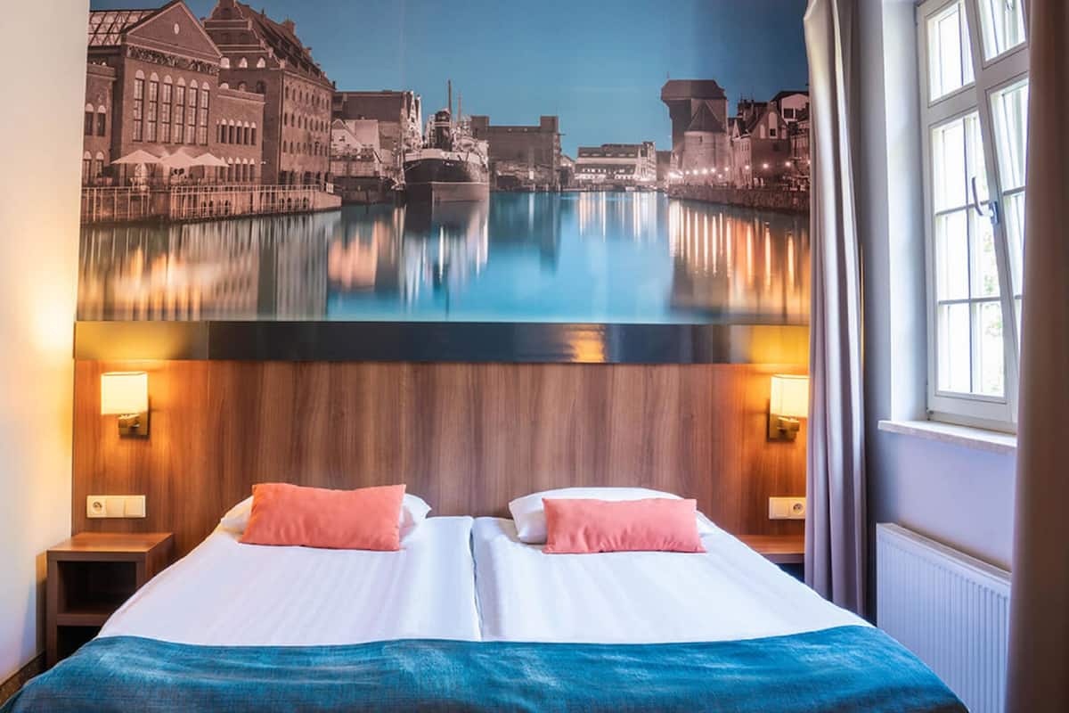Une chambre d’hôtel avec un lit, deux oreillers, une table de chevet et une fresque murale représentant un paysage de canal.